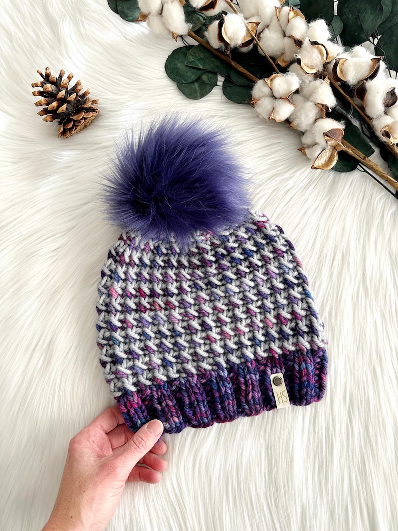 May include: A gray, purple, and pink knit beanie with a purple faux fur pom pom. The beanie has a small brown tag with the letters "HS" on it.