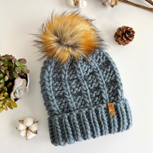 May include: A gray knitted beanie with a large, fluffy pom pom in shades of orange and brown. The beanie has a cable knit pattern and a leather tag with the initials "HS".