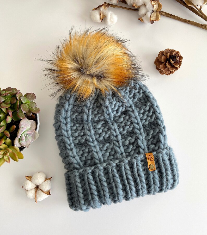 May include: A gray knitted beanie with a large, fluffy pom pom in shades of orange and brown. The beanie has a cable knit pattern and a leather tag with the initials "HS".