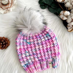 May include: A pink, blue, and white knit beanie with a fluffy white pom pom. The beanie has a small leather tag with the letters "HS" on it.