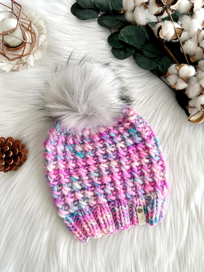 May include: A pink, blue, and white knit beanie with a fluffy white pom pom. The beanie has a small leather tag with the letters "HS" on it.