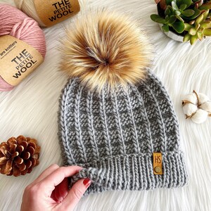 May include: Gray knitted beanie with a faux fur pom pom. The beanie has a ribbed texture and a small leather tag with the letters "HS" on it.