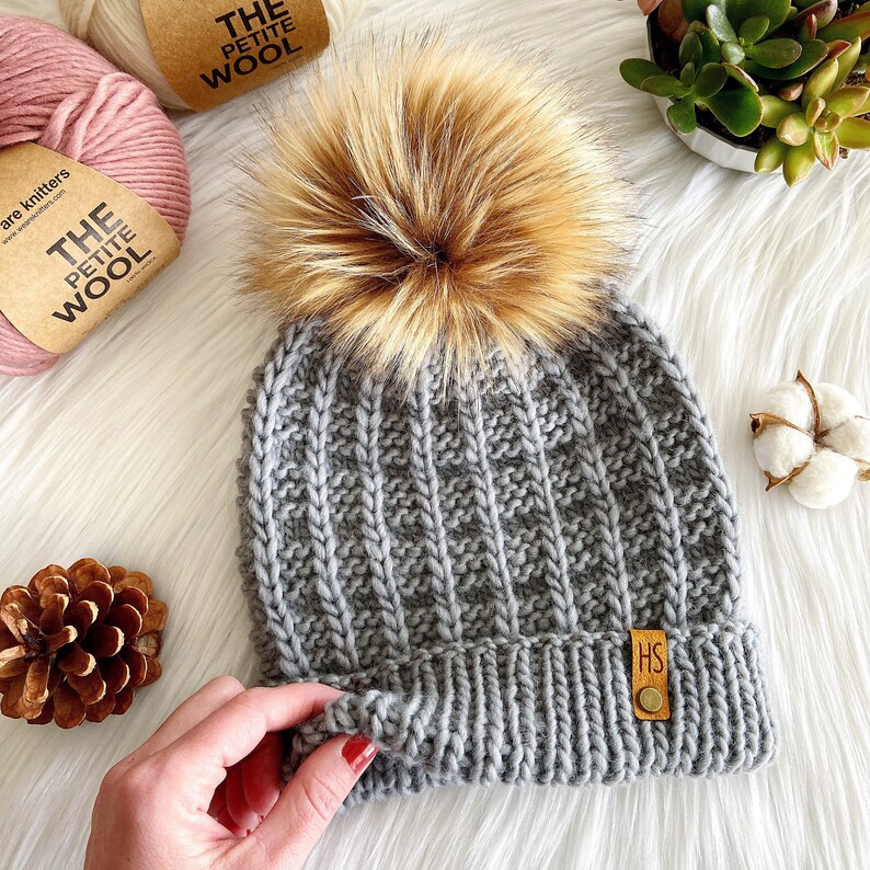 May include: Gray knitted beanie with a faux fur pom pom. The beanie has a ribbed texture and a small leather tag with the letters "HS" on it.