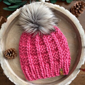 May include: A pink knitted beanie with a large faux fur pom pom. The beanie has a cable knit pattern and a brown leather tag with the initials "HS" on it.
