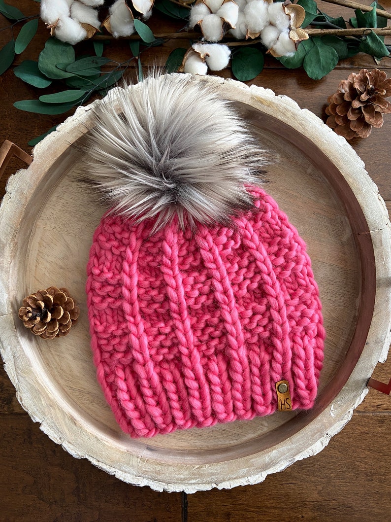 May include: A pink knitted beanie with a large faux fur pom pom. The beanie has a cable knit pattern and a brown leather tag with the initials "HS" on it.