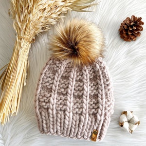 May include: A blush pink knitted beanie with a large brown faux fur pom pom. The beanie has a cable knit pattern and a brown leather tag with the letters "HS" on it.