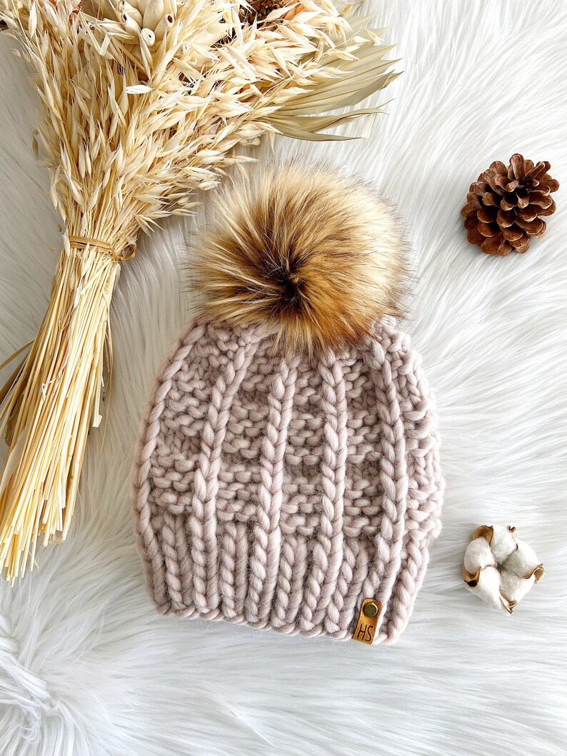 May include: A blush pink knitted beanie with a large brown faux fur pom pom. The beanie has a cable knit pattern and a brown leather tag with the letters "HS" on it.