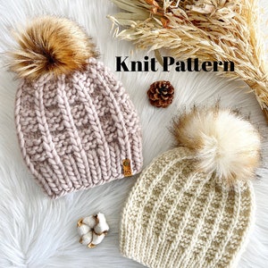 Puede incluir: Dos gorros de punto con pompones de piel sintética. Los gorros están hechos con un patrón de punto de cable. Un gorro es rosa y el otro es crema. Los gorros tienen una etiqueta de cuero con las letras "HS" en ellos. Los gorros están sobre una superficie blanca y esponjosa con flores secas y una bola de algodón.