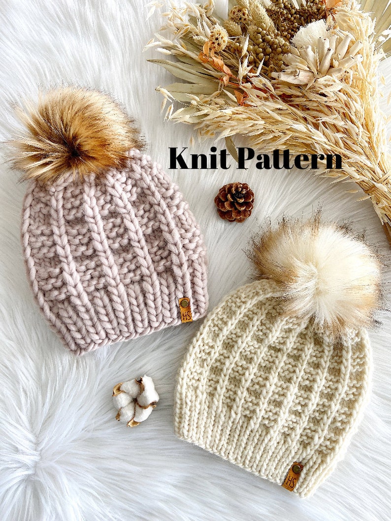 May include: Two knitted hats with faux fur pom poms. The hats are in a cable knit pattern. One hat is pink and the other is cream. The hats have a leather tag with the letters "HS" on them. The hats are on a white fluffy surface with dried flowers and a cotton boll.