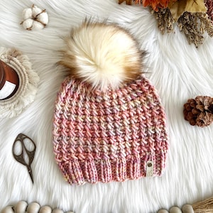 May include: A pink, yellow, and white knit beanie with a fluffy brown and white pom pom. The beanie has a small brown leather tag with the letters "HS" on it.