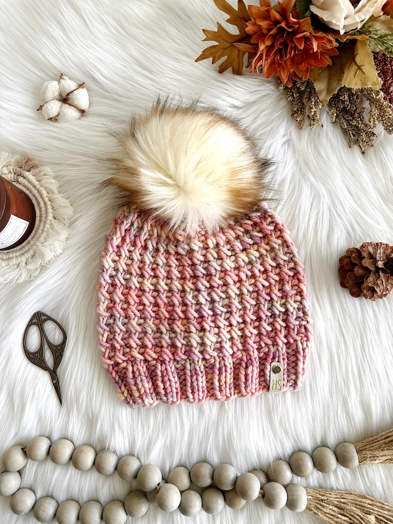 May include: A pink, yellow, and white knit beanie with a fluffy brown and white pom pom. The beanie has a small brown leather tag with the letters "HS" on it.