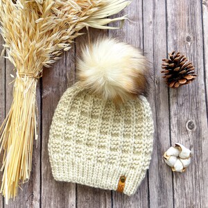 May include: A cream-colored knitted beanie with a fluffy faux fur pom pom. The beanie has a textured knit pattern and a brown leather tag with the letters "HS" on it.