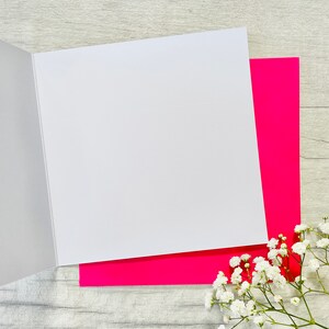 May include: A blank white card with a pink envelope. The card is folded in half and the envelope is open. The card and envelope are on a light wood surface.