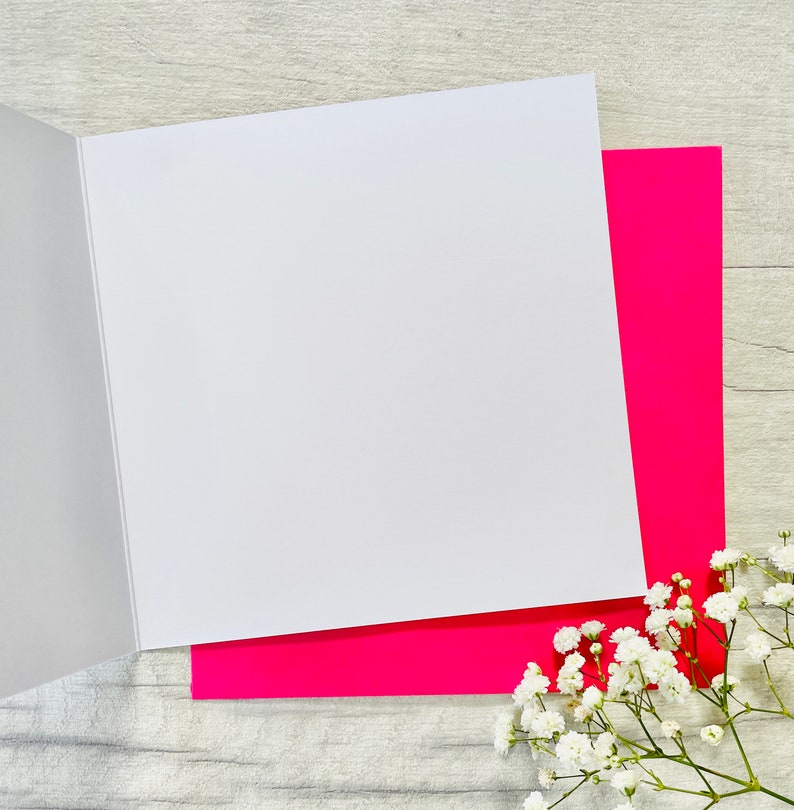 May include: A blank white card with a pink envelope. The card is folded in half and the envelope is open. The card and envelope are on a light wood surface.
