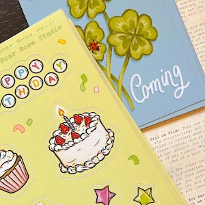May include: A light yellow sticker sheet featuring a birthday cake, a cupcake, a cat in a party hat, and colorful stars, with the text "HAPPY BIRTHDAY" and "Scar Rose Studio". Behind it, a blue card displays green four-leaf clovers and ladybugs.