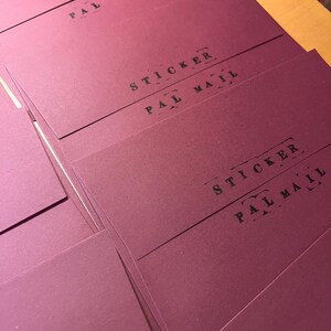 May include: A stack of rectangular, deep burgundy paper envelopes. Each envelope features the black printed text STICKER and PAL MAIL, with the word STICKER underlined by a dashed line and PAL MAIL framed by small decorative brackets.