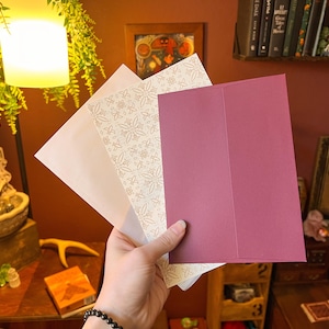 May include: A set of three rectangular envelopes held together. The front envelope is solid deep magenta, the middle envelope features a white and gold floral pattern, and the back envelope is plain white. Each envelope has a pointed flap design.