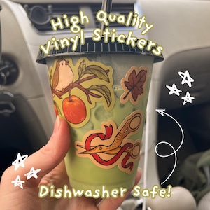 May include: A clear plastic cup filled with a light green beverage, featuring a black lid and straw. The cup is decorated with vinyl stickers of a bird on a branch with an apple, a brown leaf, and scissors with a red ribbon. Text reads High Quality Vinyl Stickers and Dishwasher Safe!