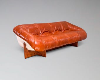 May include: A mid-century modern sofa with a tufted, orange-brown leather seat. The sofa has a white base and rests on a dark wood frame. The design is a classic example of vintage furniture.