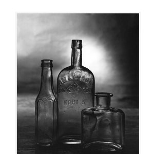 May include: Black and white photograph of three antique glass bottles. The bottles vary in shape and size, with one tall and slender, another rounded with embossed text, and a third, a small square bottle. The bottles are arranged on a dark surface.