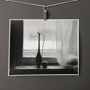 May include: A black and white photograph of a vase and a sphere sitting on a windowsill. The vase is tall and slender with a rounded base. The sphere is small and smooth. The window is covered in condensation.