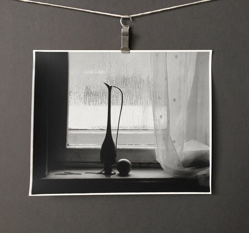 May include: A black and white photograph of a vase and a sphere sitting on a windowsill. The vase is tall and slender with a rounded base. The sphere is small and smooth. The window is covered in condensation.