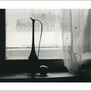 May include: A black and white photo of a tall, slender vase with a long, thin neck and a round, bulbous base. The vase is sitting on a windowsill, with a sheer curtain behind it. The window is covered in condensation, and the light from outside is casting a soft glow on the scene.