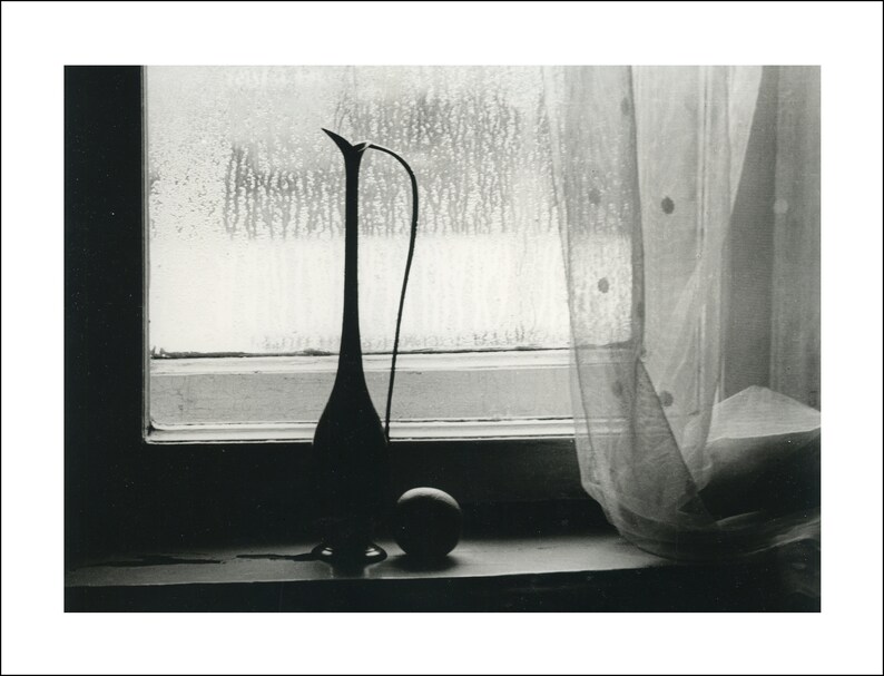 May include: A black and white photo of a tall, slender vase with a long, thin neck and a round, bulbous base. The vase is sitting on a windowsill, with a sheer curtain behind it. The window is covered in condensation, and the light from outside is casting a soft glow on the scene.