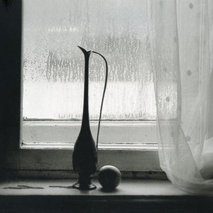 May include: A black and silver vase with a long neck and a round body sits on a windowsill. A single, round, light-colored fruit sits next to the vase. A sheer white curtain hangs in the background.