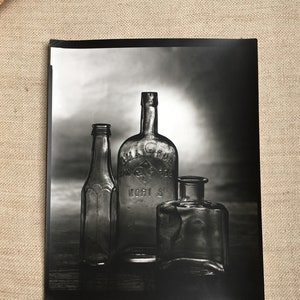 May include: A black and white photograph of three antique glass bottles. The bottles vary in shape and size, with one taller and more ornate than the others. The middle bottle has text on it. The image is lit from behind, creating shadows.