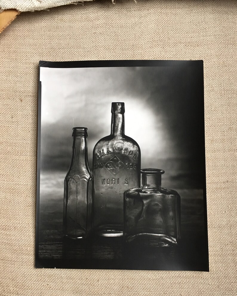 May include: A black and white photograph of three antique glass bottles. The bottles vary in shape and size, with one taller and more ornate than the others. The middle bottle has text on it. The image is lit from behind, creating shadows.