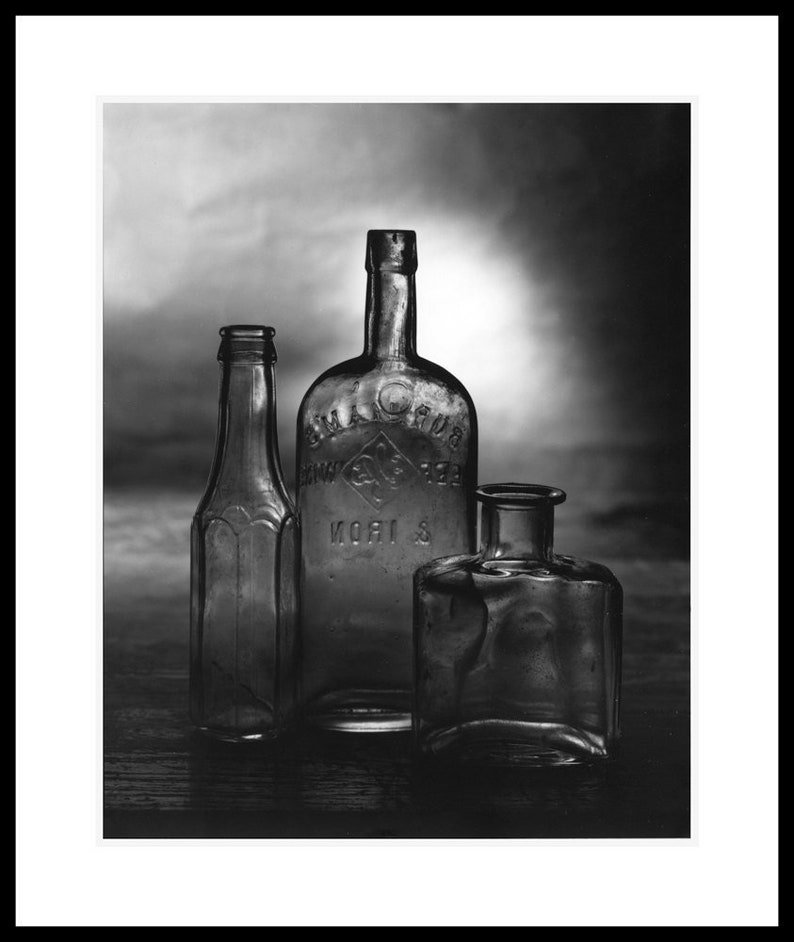 May include: Black and white photograph of three antique glass bottles. The bottles vary in shape and size, with one tall and slender, another rounded with embossed text, and a third, a small square bottle. The bottles are arranged on a dark surface.