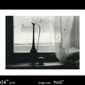 May include: A black and white photograph of a still life featuring a ceramic vase with a long neck and a round, smooth object on a windowsill. The vase is silhouetted against a window with a sheer curtain.