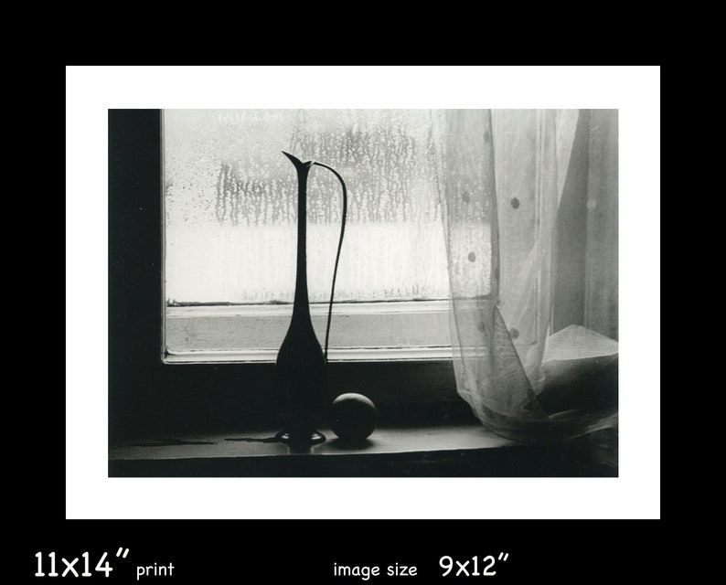 May include: A black and white photograph of a still life featuring a ceramic vase with a long neck and a round, smooth object on a windowsill. The vase is silhouetted against a window with a sheer curtain.