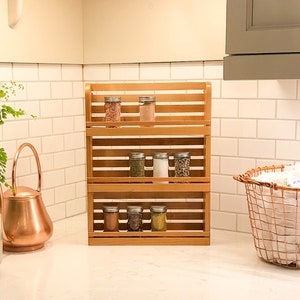 May include: A three-tiered wooden spice rack filled with glass jars of spices. The rack is made of light-colored wood and sits on a white countertop. The background includes a white tiled backsplash and a copper watering can.