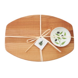 May include: A wooden serving board with a small, round dish. The dish features a green olive branch design with black olives. The set is tied with natural twine and includes a tag with a pine tree illustration.