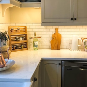 May include: A white kitchen countertop with a wooden spice rack, a bottle of wine, two wine glasses, two wooden cutting boards, a white pitcher, a wicker basket, and a stainless steel dishwasher.
