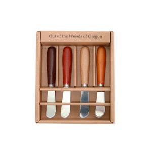 May include: Set of five wooden handled butter knives in a cardboard box. The box is labeled "Out of the Woods of Oregon". The knives have different colored wooden handles: brown, red, natural wood, and orange.