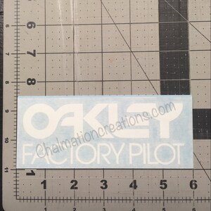 May include: White vinyl sticker with the text "OAKLEY FACTORY PILOT" and the website "chalmationcreations.com" printed in white.