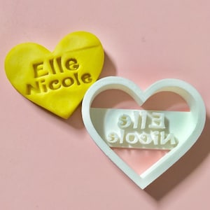 May include: A white heart-shaped cookie cutter with the words "Elle Nicole" embossed on it. The cutter is next to a yellow heart-shaped cookie with the same words imprinted on it.