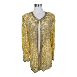Pode incluir: Uma jaqueta longa e aberta na frente, em um tom amarelo dourado, adornada com renda floral intrincada e embelezada com contas de prata. A jaqueta apresenta mangas compridas e decote em V, exibida em um manequim.
