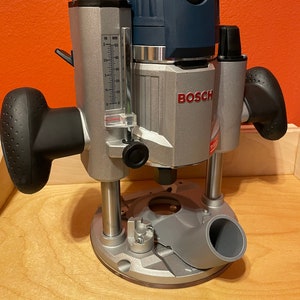 May include: A blue and silver Bosch router with a depth gauge and a dust collection port. The router is on a wooden surface.