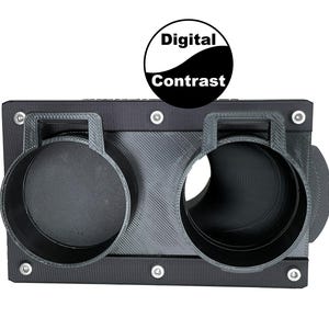May include: A black, rectangular device with two circular openings and a textured surface. The device has a handle above each opening and is secured with silver screws. The text "Digital Contrast" is displayed above the device.