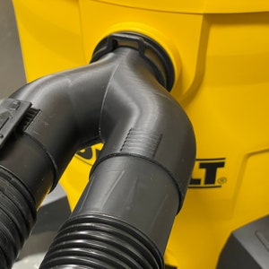 May include: A black plastic Y-shaped vacuum hose attachment with a locking mechanism, connected to a yellow vacuum cleaner. The attachment has ribbed detailing and is designed to split the vacuum's suction.