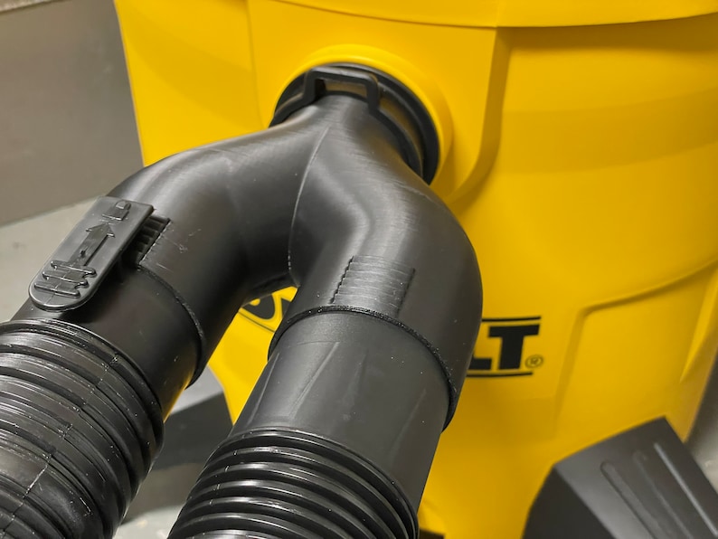 May include: A black plastic Y-shaped vacuum hose attachment with a locking mechanism, connected to a yellow vacuum cleaner. The attachment has ribbed detailing and is designed to split the vacuum's suction.