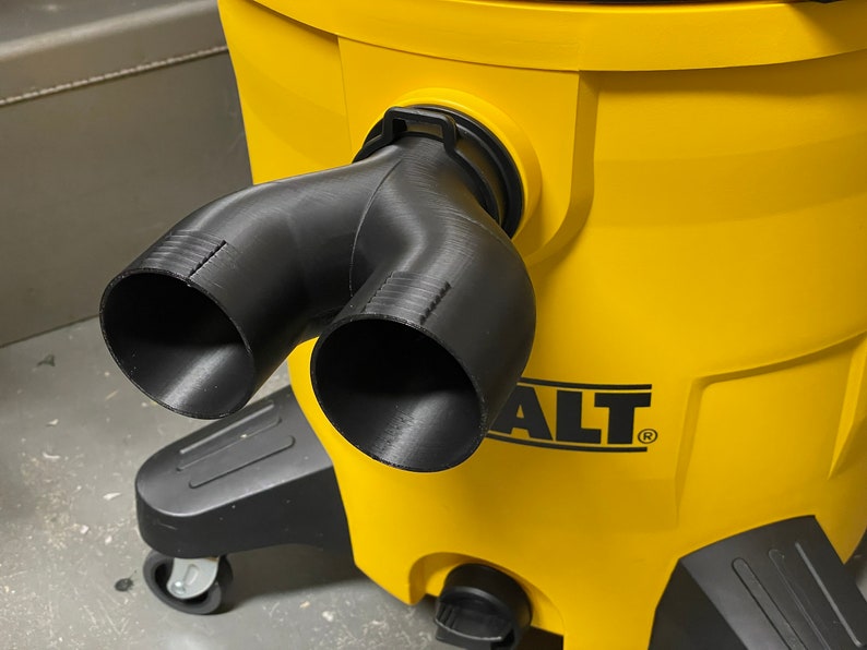 May include: A black, Y-shaped vacuum hose attachment is connected to a bright yellow DEWALT vacuum cleaner. The attachment has two openings and is made of a 3D-printed material. The vacuum cleaner has a black base with wheels.