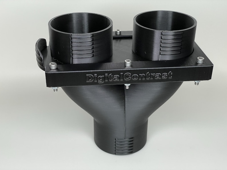 May include: Black 3D-printed plastic component with two cylindrical openings and a funnel-shaped base. The component has a rectangular top with the text "DigitalContrast" and is secured with screws. The design suggests a functional, possibly industrial, product.