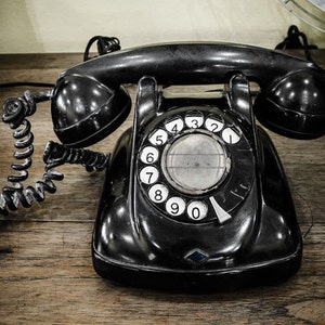 May include: A black rotary dial phone with a coiled cord. The phone has a black dial with white numbers 0-9. The phone is sitting on a wooden surface.