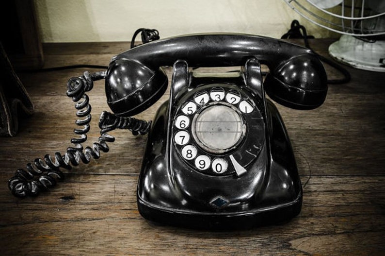 May include: A black rotary dial phone with a coiled cord. The phone has a black dial with white numbers 0-9. The phone is sitting on a wooden surface.
