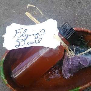 May include: A small brown glass bottle with a black cap, labeled "Flying Devil", sitting in a terracotta bowl with a purple amethyst crystal.